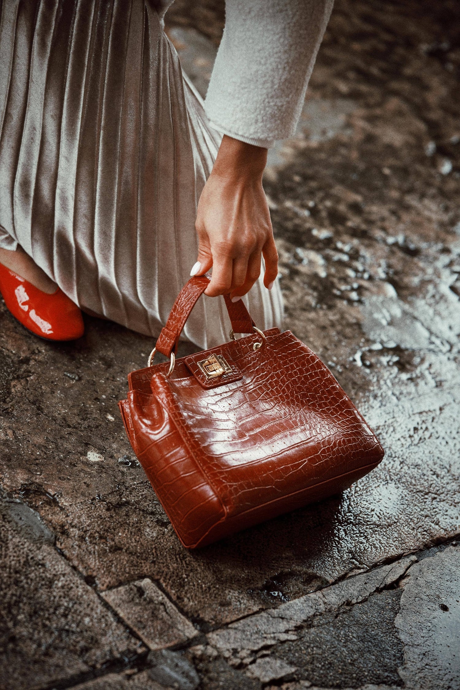 Femme tenant un sac à main marron effet crocodile posé au sol dans une tenue élégante.